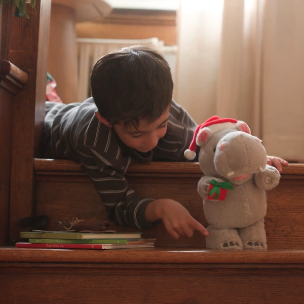 A small, dark-haired boy of about seven years old lays at the top of a set of wooden stairs in his pajamas; sitting on the step directly below him is a musical dancing Christmas hippo plush toy.